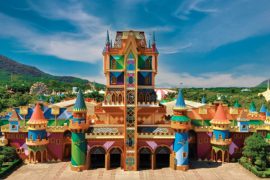 Fotografia colorida do castelo do parque temático Beto Carrero World, localizado em Penha, Santa Catarina. O castelo apresenta uma arquitetura vibrante e lúdica, com torres pontiagudas em tons de azul, vermelho e amarelo, além de vitrais coloridos no centro. Ao fundo, é possível ver a vegetação e o céu azul com algumas nuvens. O design do castelo remete a um estilo mágico e encantado, ideal para o ambiente de um parque de diversões.