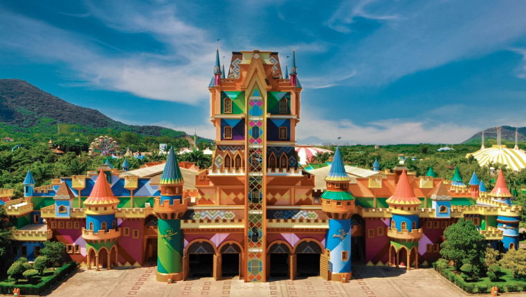 Fotografia colorida do castelo do parque temático Beto Carrero World, localizado em Penha, Santa Catarina. O castelo apresenta uma arquitetura vibrante e lúdica, com torres pontiagudas em tons de azul, vermelho e amarelo, além de vitrais coloridos no centro. Ao fundo, é possível ver a vegetação e o céu azul com algumas nuvens. O design do castelo remete a um estilo mágico e encantado, ideal para o ambiente de um parque de diversões.