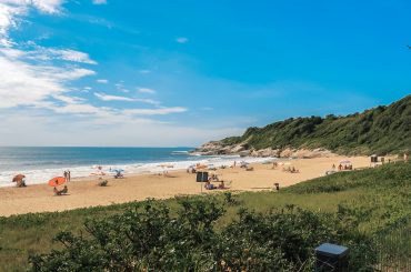 Imagem de uma praia tranquila com areia dourada e águas azuladas em um dia ensolarado. Algumas pessoas estão aproveitando o local com guarda-sóis coloridos espalhados pela areia. No lado direito, há uma área com vegetação verdejante e formações rochosas que complementam o cenário natural. O céu está claro e limpo, reforçando a atmosfera relaxante e convidativa.