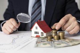 Imagem de um homem em traje formal, segurando uma lupa próxima a uma miniatura de uma casa com telhado vermelho. Ao lado da casa, há pilhas de moedas e cédulas de dinheiro, indicando análise financeira ou investimentos imobiliários. Sobre a mesa, também é possível ver gráficos que sugerem estudo de mercado ou planejamento financeiro.