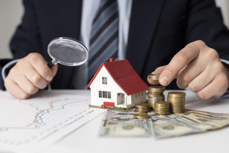 Imagem de um homem em traje formal, segurando uma lupa próxima a uma miniatura de uma casa com telhado vermelho. Ao lado da casa, há pilhas de moedas e cédulas de dinheiro, indicando análise financeira ou investimentos imobiliários. Sobre a mesa, também é possível ver gráficos que sugerem estudo de mercado ou planejamento financeiro.