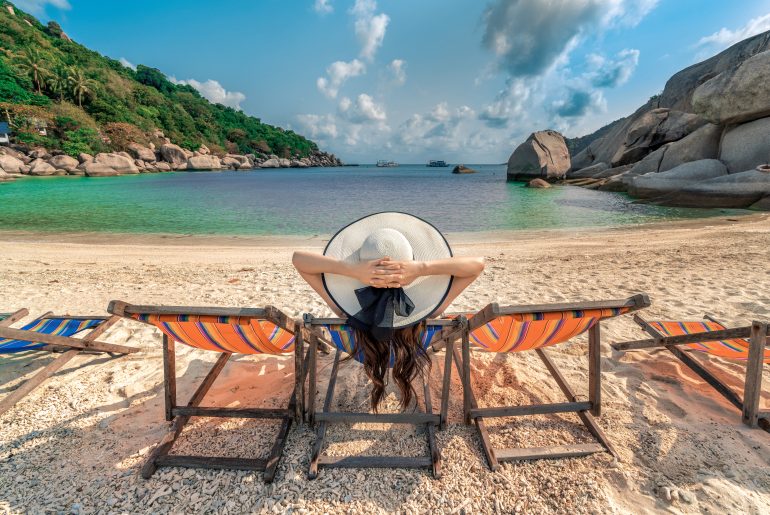 Uma mulher relaxa em uma cadeira de praia colorida, virada de costas, usando um chapéu branco de aba larga enquanto aprecia a paisagem de uma praia paradisíaca. A areia clara e fina contrasta com a água cristalina, que varia em tons de azul e verde. Rochas grandes e vegetação tropical cercam a enseada, criando um cenário tranquilo e encantador, com um céu azul pontuado por algumas nuvens. Ao fundo, é possível avistar pequenos barcos ancorados no mar calmo.