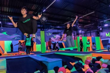 Uma imagem de um parque indoor de trampolins, com pessoas pulando e se divertindo em um ambiente colorido e iluminado por luzes neon. No centro, duas pessoas estão em destaque, saltando com expressão animada. Ao fundo, é possível ver outras pessoas utilizando os trampolins e a área com blocos de espuma, em um espaço vibrante com decoração temática que inclui desenhos e elementos gráficos nas paredes.