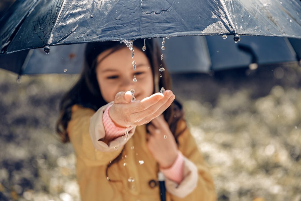 O Que Fazer em Balneário Camboriú com Chuva? Uma imagem de uma criança sorrindo e segurando um guarda-chuva azul escuro enquanto gotas de chuva escorrem pela borda. Ela estende a mão para sentir as gotas caindo, vestindo uma capa de chuva amarela com detalhes em rosa. O fundo está desfocado, mostrando um ambiente externo, provavelmente um parque ou área arborizada.