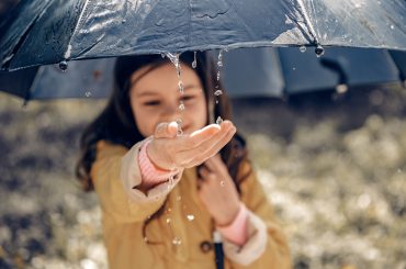Uma imagem de uma criança sorrindo e segurando um guarda-chuva azul escuro enquanto gotas de chuva escorrem pela borda. Ela estende a mão para sentir as gotas caindo, vestindo uma capa de chuva amarela com detalhes em rosa. O fundo está desfocado, mostrando um ambiente externo, provavelmente um parque ou área arborizada.