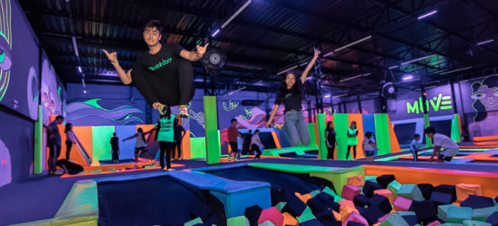 Uma imagem de um parque indoor de trampolins, com pessoas pulando e se divertindo em um ambiente colorido e iluminado por luzes neon. No centro, duas pessoas estão em destaque, saltando com expressão animada. Ao fundo, é possível ver outras pessoas utilizando os trampolins e a área com blocos de espuma, em um espaço vibrante com decoração temática que inclui desenhos e elementos gráficos nas paredes.