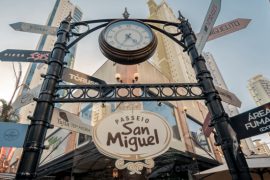 Imagem do portal de entrada do Passeio San Miguel, em Balneário Camboriú. O portal é decorado com um relógio clássico no topo e placas direcionais indicando diferentes locais e estabelecimentos, como restaurantes e áreas específicas. O ambiente é cercado por prédios altos e árvores, com um céu limpo ao fundo, transmitindo a atmosfera charmosa e urbana do local.