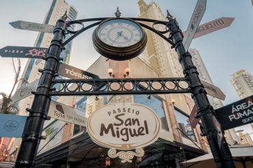 Imagem do portal de entrada do Passeio San Miguel, em Balneário Camboriú. O portal é decorado com um relógio clássico no topo e placas direcionais indicando diferentes locais e estabelecimentos, como restaurantes e áreas específicas. O ambiente é cercado por prédios altos e árvores, com um céu limpo ao fundo, transmitindo a atmosfera charmosa e urbana do local.
