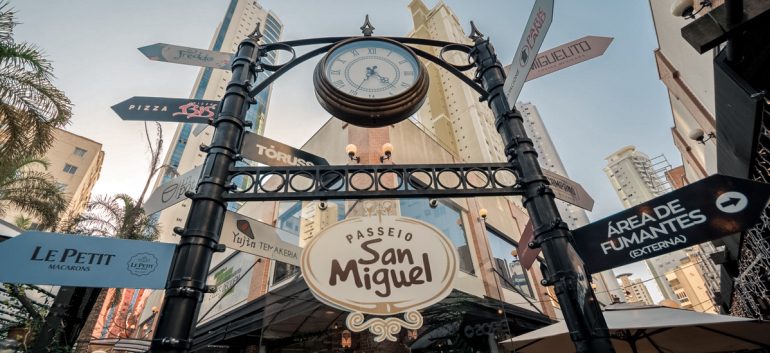 Imagem do portal de entrada do Passeio San Miguel, em Balneário Camboriú. O portal é decorado com um relógio clássico no topo e placas direcionais indicando diferentes locais e estabelecimentos, como restaurantes e áreas específicas. O ambiente é cercado por prédios altos e árvores, com um céu limpo ao fundo, transmitindo a atmosfera charmosa e urbana do local.