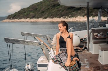 Uma mulher sentada na borda de uma lancha, vestindo maiô preto e saia estampada, sorrindo enquanto observa a paisagem ao seu redor. A lancha está ancorada em um local tranquilo, com água azul calma e uma área costeira ao fundo, repleta de vegetação verde e rochas. O deck da lancha exibe detalhes de luxo, como assentos e utensílios de uso pessoal, sugerindo um passeio relaxante.