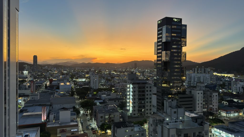 Vista panorâmica de uma cidade ao entardecer, com o céu iluminado por tons alaranjados e azulados. Prédios altos, incluindo um edifício moderno identificado como 'Sky', destacam-se no cenário urbano iluminado, enquanto montanhas ao fundo criam um contraste natural. A imagem transmite a combinação de vida urbana com a beleza natural.
