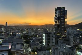 Vista panorâmica de uma cidade ao entardecer, com o céu iluminado por tons alaranjados e azulados. Prédios altos, incluindo um edifício moderno identificado como 'Sky', destacam-se no cenário urbano iluminado, enquanto montanhas ao fundo criam um contraste natural. A imagem transmite a combinação de vida urbana com a beleza natural.