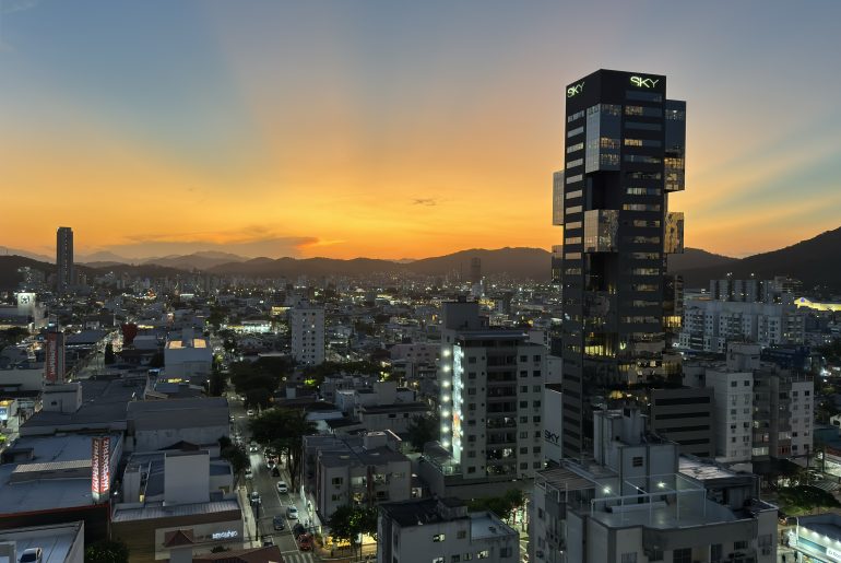 Vista panorâmica de uma cidade ao entardecer, com o céu iluminado por tons alaranjados e azulados. Prédios altos, incluindo um edifício moderno identificado como 'Sky', destacam-se no cenário urbano iluminado, enquanto montanhas ao fundo criam um contraste natural. A imagem transmite a combinação de vida urbana com a beleza natural.