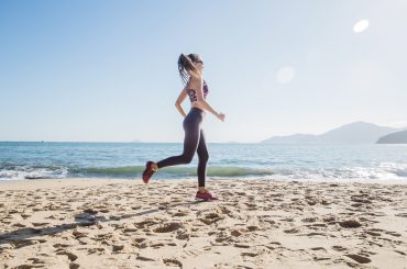 Mulher correndo na areia de uma praia em um dia ensolarado, com o mar azul ao fundo e montanhas na linha do horizonte, transmitindo energia e estilo de vida saudável