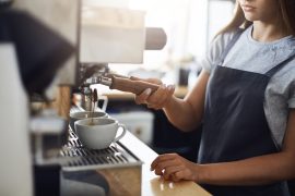 Barista preparando um café espresso em uma máquina profissional, com duas xícaras brancas posicionadas para receber a bebida. O ambiente é iluminado e acolhedor, ideal para apreciar um bom café.