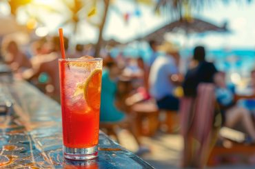 Drink colorido servido em um copo alto com gelo e uma fatia de laranja, em um bar ao ar livre na praia, com pessoas descontraídas e um cenário ensolarado ao fundo.
