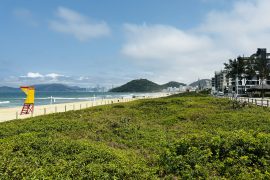 Vista da Praia Brava em um dia ensolarado, com areia clara, vegetação costeira, um posto de guarda-vidas colorido e edifícios modernos ao longo da orla. Ao fundo, montanhas e o mar azul complementam a paisagem,