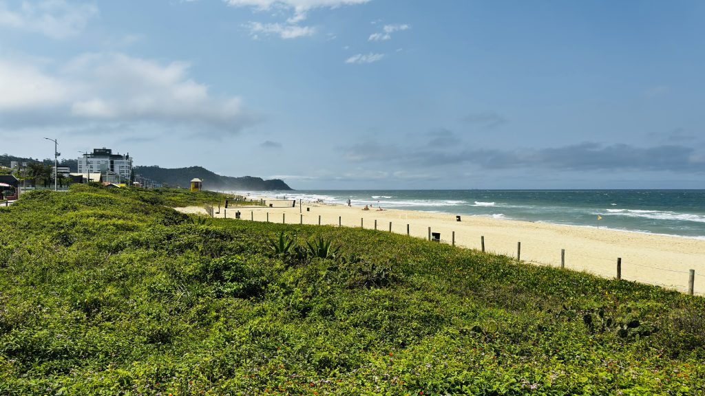 Vista panorâmica de uma praia com faixa de areia dourada e mar azul, cercada por vegetação verde e algumas construções ao fundo, em um dia ensolarado com poucas nuvens.