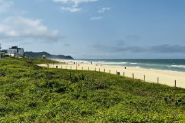 Vista panorâmica de uma praia com faixa de areia dourada e mar azul, cercada por vegetação verde e algumas construções ao fundo, em um dia ensolarado com poucas nuvens.