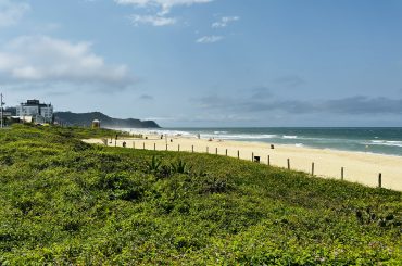 Vista panorâmica de uma praia com faixa de areia dourada e mar azul, cercada por vegetação verde e algumas construções ao fundo, em um dia ensolarado com poucas nuvens.